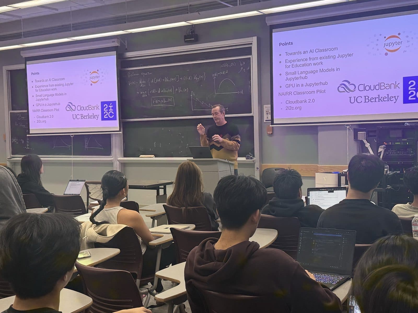 A guest lecturer speaking to DeCal students in a Berkeley lecture hall, slides projected on two screens behind him.