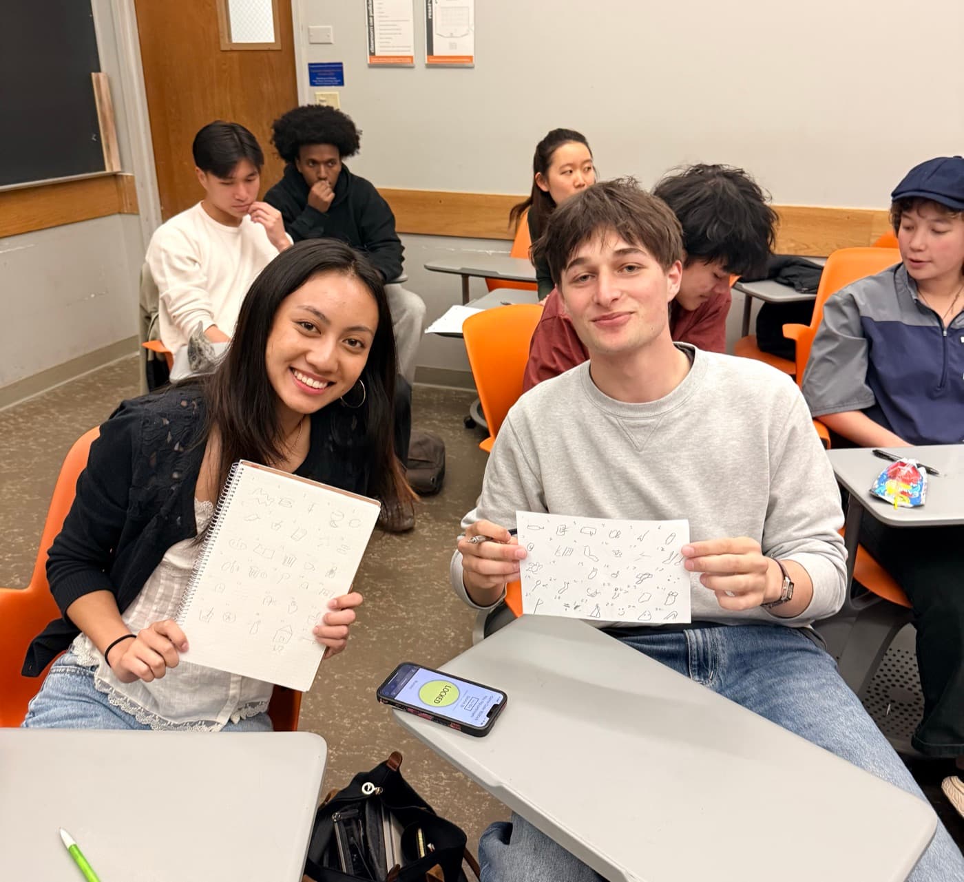 Two committee members holding up sheets of paper covered in quick sketches during a drawing game in class
