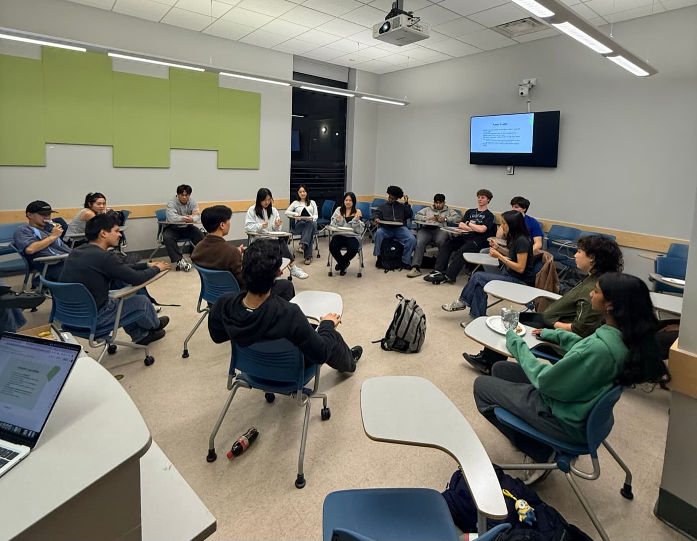 Committee members seated in a wide circle of desk chairs in a classroom, talking with a presentation on the screen behind them